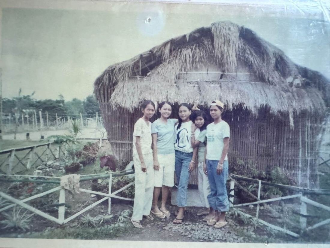 An old photograph of five young people standing in front of a thatched hut on the school grounds